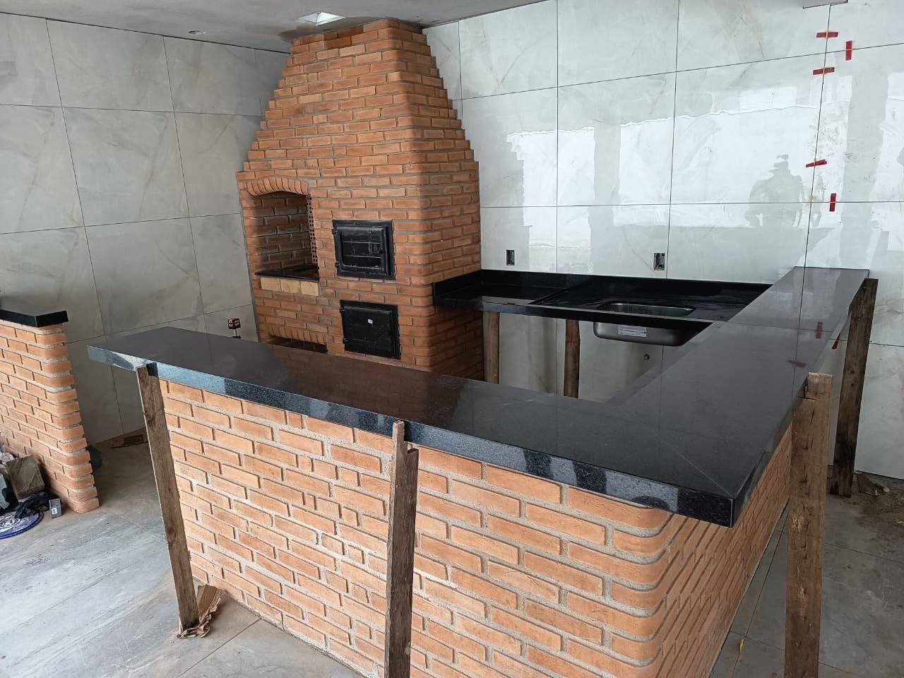 Área gourmet com churrasqueira de tijolos e bancada em L em granito preto polido com cuba embutida
