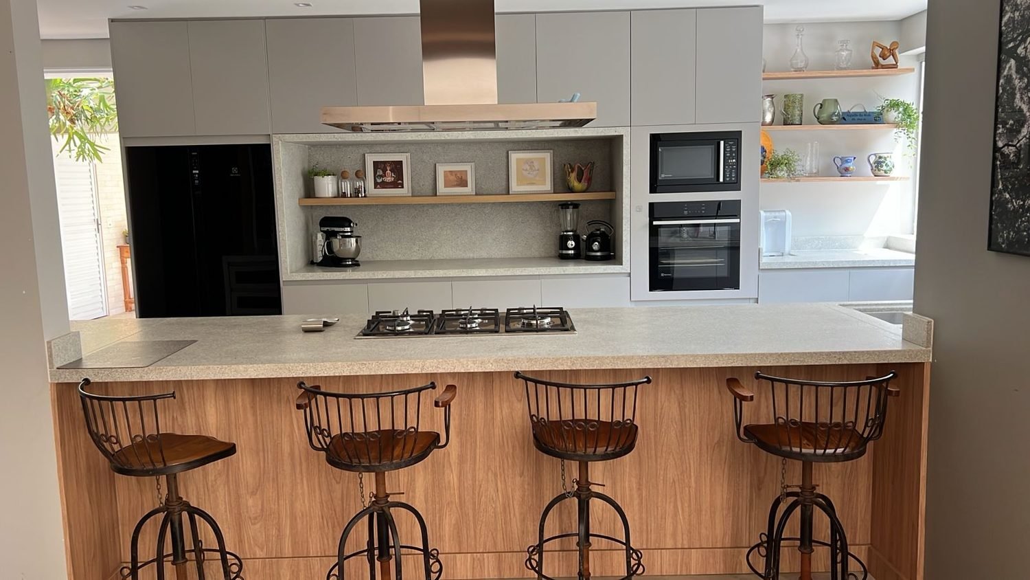 Cozinha com bancada em granito cinza, cooktop, nicho revestido em pedra e balcão com banquetas